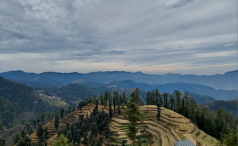 A breathtaking view of the mountain range in Himachal Pradesh, India, with stunning clouds hovering above and a stepped farm visible in the foreground. The majestic mountains stand tall, their peaks partially obscured by the clouds, while lush greenery adorns the lower slopes. The stepped farm adds to the scenic beauty, showcasing human interaction with the natural landscape in a harmonious manner.