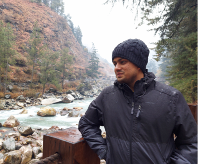 A photograph of an individual in Kasol, with a river in the background and mist-covered mountains. The person is wearing a jacket and leaning on a wooden pillar of a fence, with their elbow resting on it. The serene river flows in the background, and mist partially shrouds the towering mountains, creating a picturesque scene of tranquility and natural beauty.