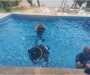 A photograph of an individual in a pool in Phuket, Thailand, engaged in a scuba diving course. The person is fully geared up in scuba diving equipment, with their hands positioned on their dive mask. They are submerged in the pool, surrounded by clear blue water. The scene captures the excitement and focus of learning to scuba dive in a tropical setting.